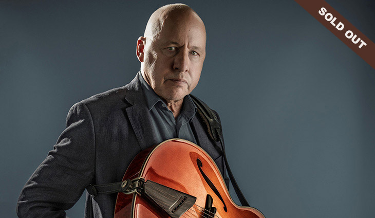 Mark Knopfler holding a guitar with a sold out banner